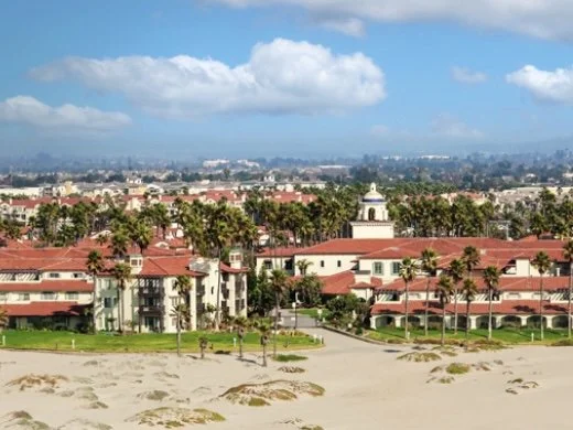 Beach with sandy shoreline in the foreground, with buildings and palm trees in the background under a partly cloudy sky.