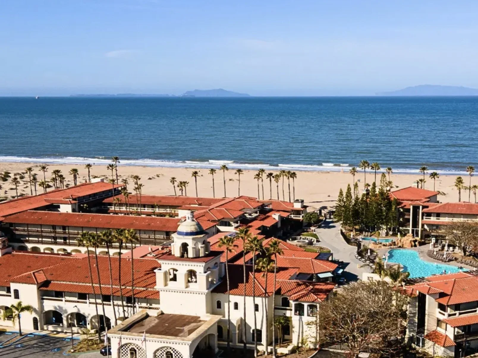 Aerial view of a beachside resort with red-tiled roofs, palm trees, a sandy beach, and the ocean in the background.