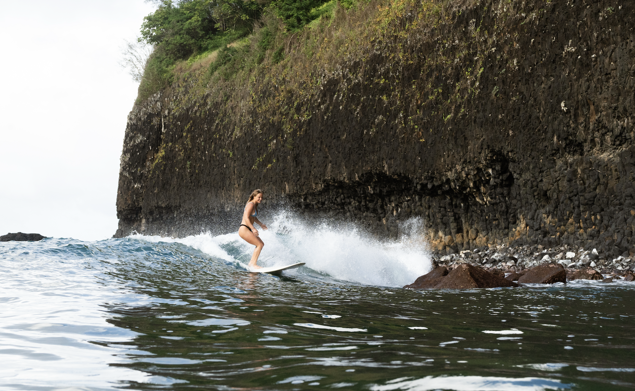 A woman surfing on a wave near a rocky coastline with a steep cliff covered in green vegetation.