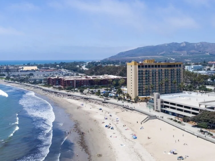 Aerial view of a beach with umbrellas, hotels, and a road with cars, with mountains in the background and clear blue sky.