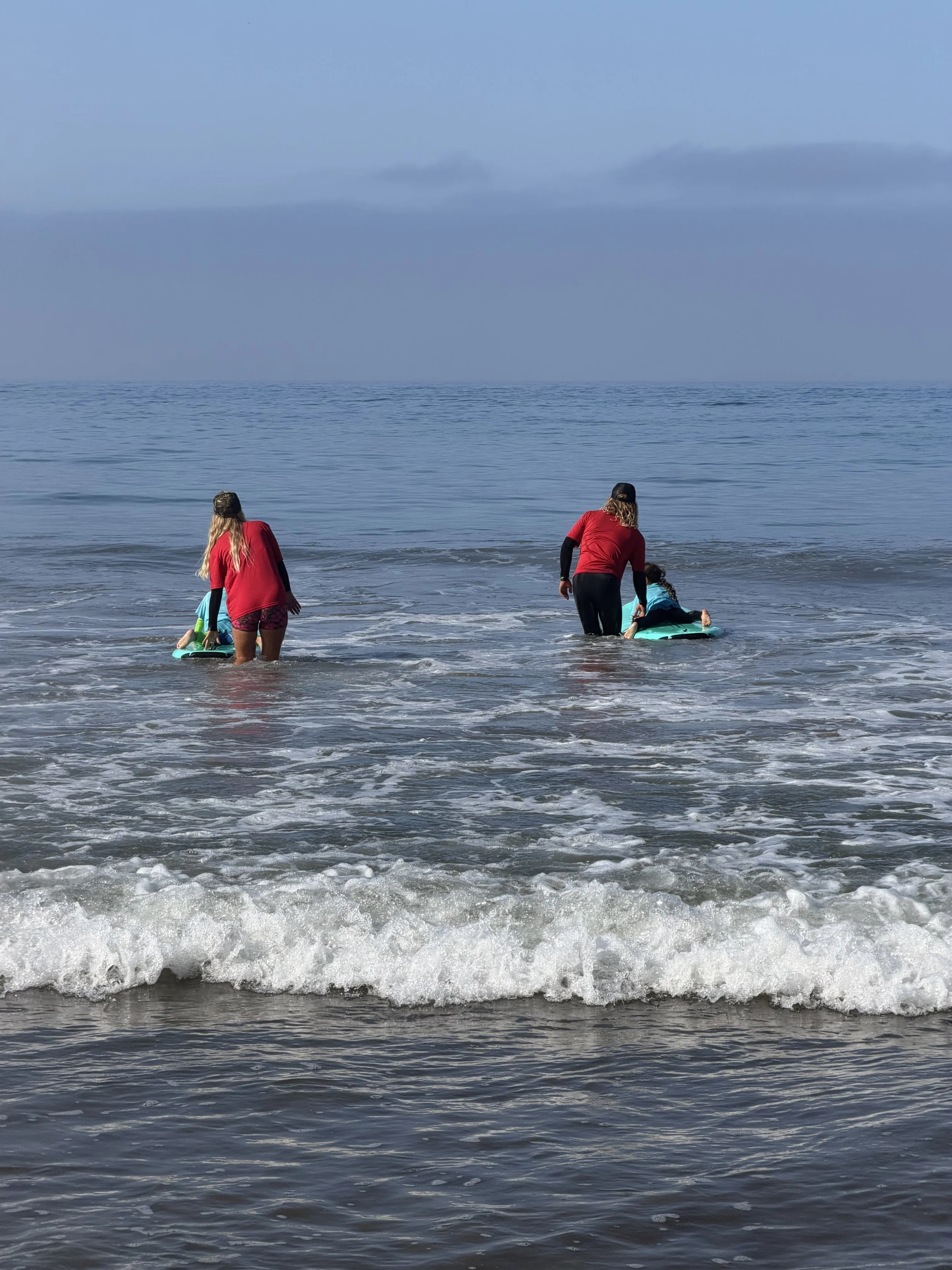 Two children and two adults at the beach, with one adult assisting a child to stand on a surfboard in the shallow water, at a beach with a calm ocean and cloudy sky.