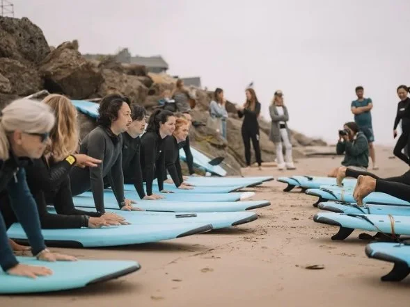 Group of people preparing surfboards on a sandy beach with rocks, some standing and others kneeling, while others observe or take photos.