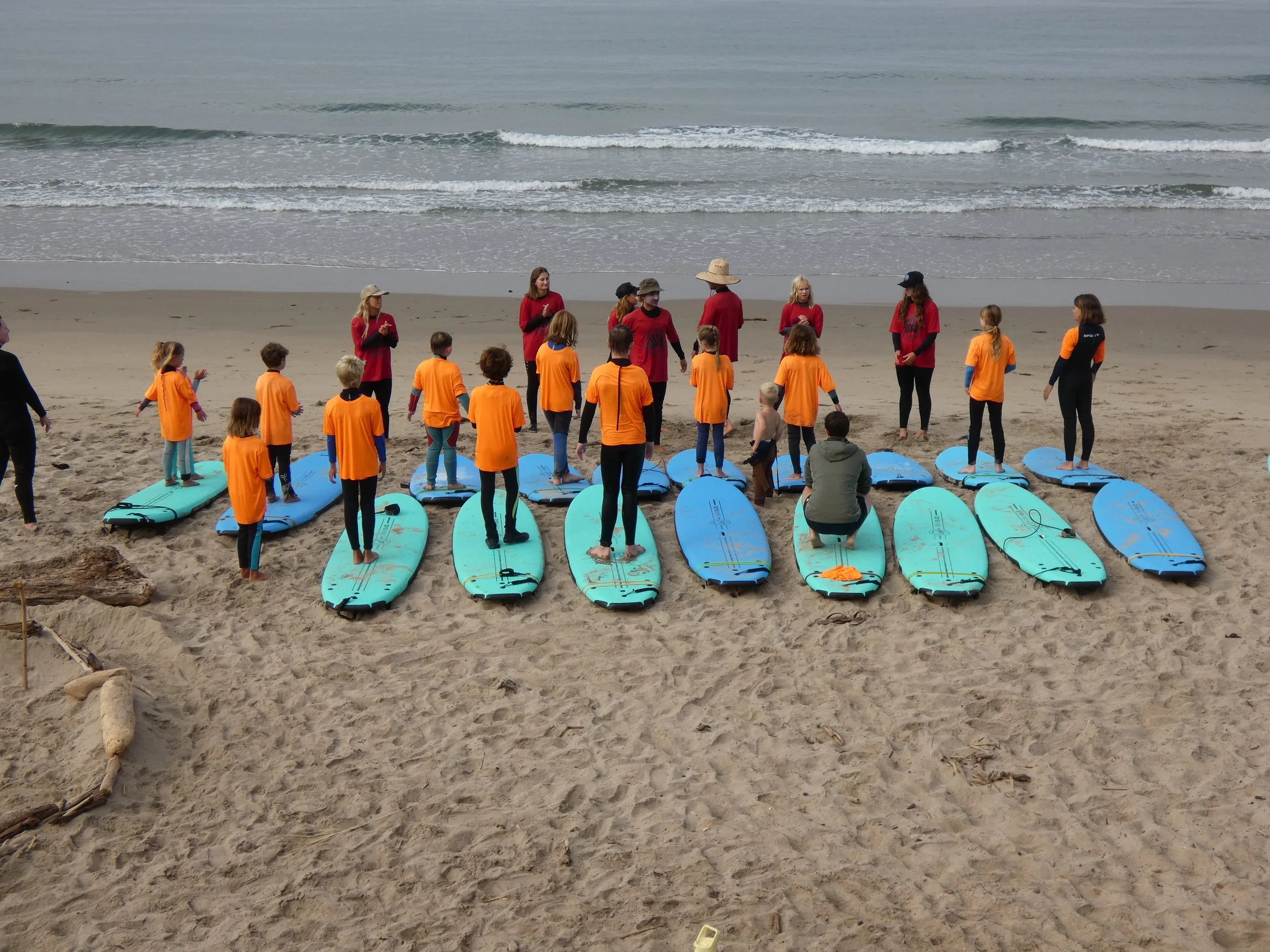 Children and adults gathered on a sandy beach with surfboards, with the ocean and waves in the background, participating in a surf lesson or group activity.