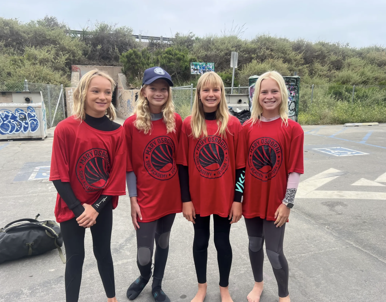 Four young girls standing together outdoors, wearing red Mary Osborne surf academy shirts, with crash pads and a graffiti-covered fence nearby.