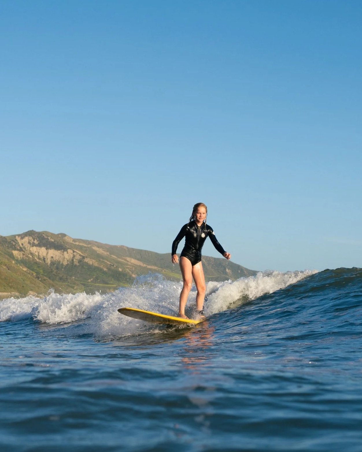 A young girl wearing a black wetsuit surfing on a yellow surfboard on the ocean with hills in the background and a clear blue sky.