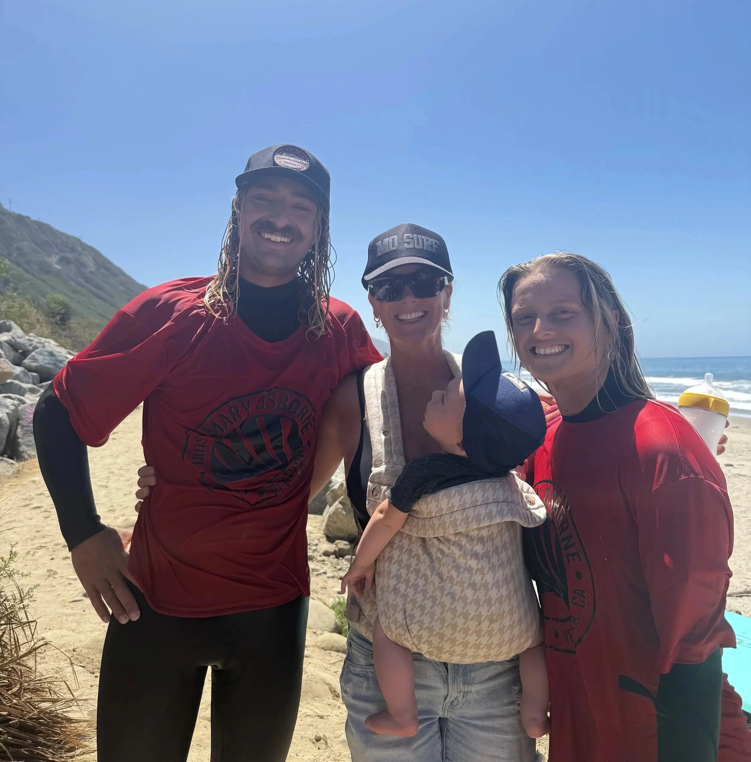 Three smiling women and a man outdoors near the beach, with a hill in the background and clear blue sky, taking a group photo.