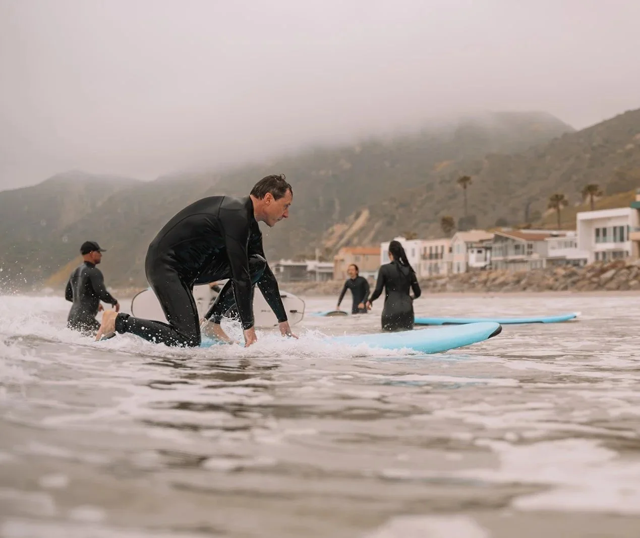 People in wetsuits and helmets preparing to surf on a cloudy day at the beach with houses and hills in the background.