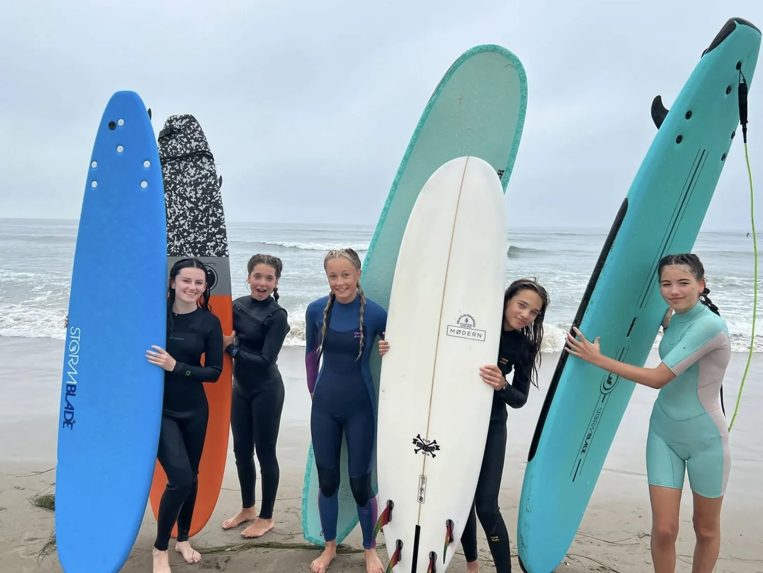 Group of five girls standing on the beach holding surfboards, smiling, with the ocean and cloudy sky in the background.