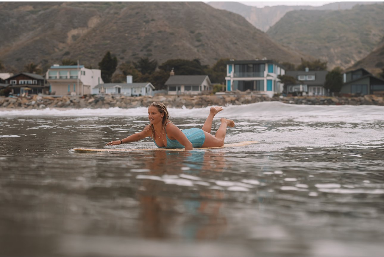 Woman lying on a surfboard in shallow ocean water near the beach with houses and mountains in the background.