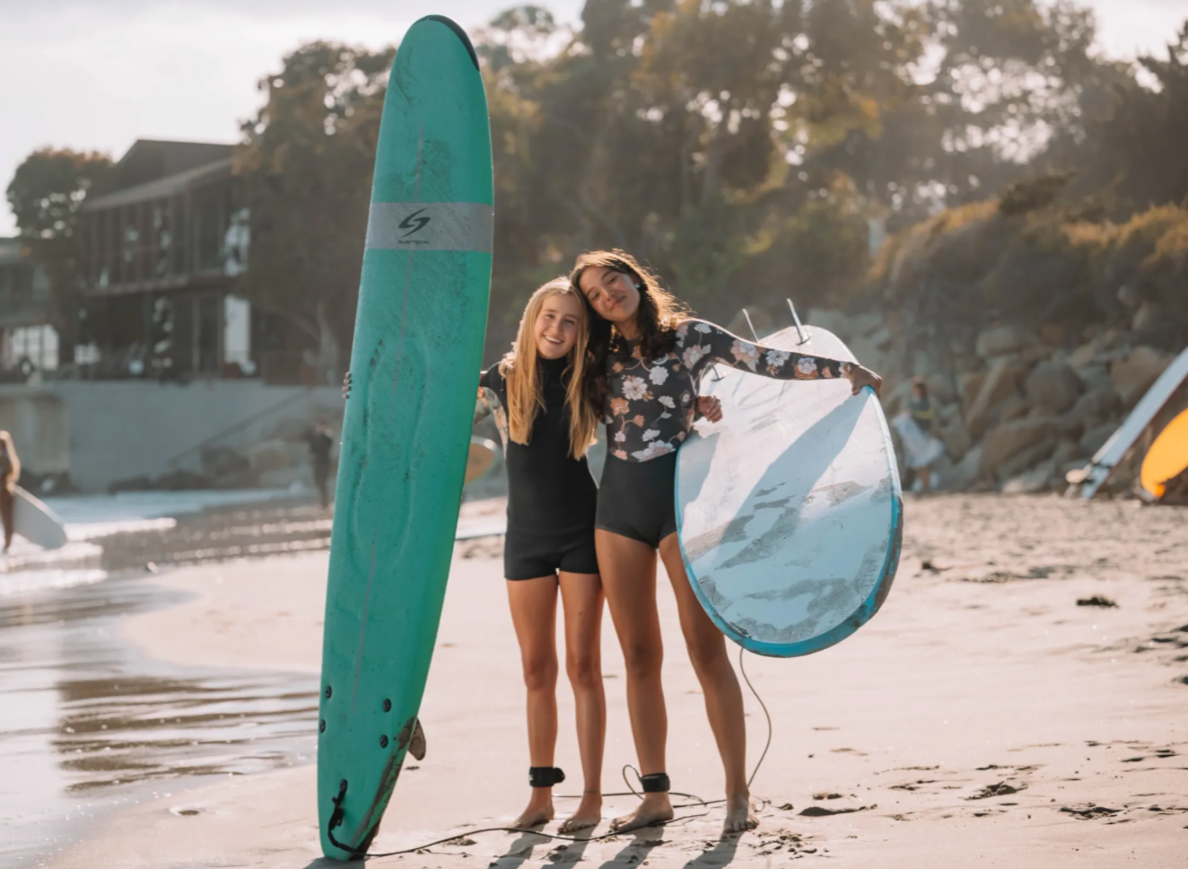 Two young women standing on a sandy beach holding surfboards and smiling, with a house and trees in the background during sunset.