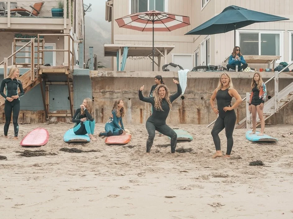 Group of women and kids on a sandy beach with surfboards, some sitting, standing, or laughing, while others relax under umbrellas near beach houses.