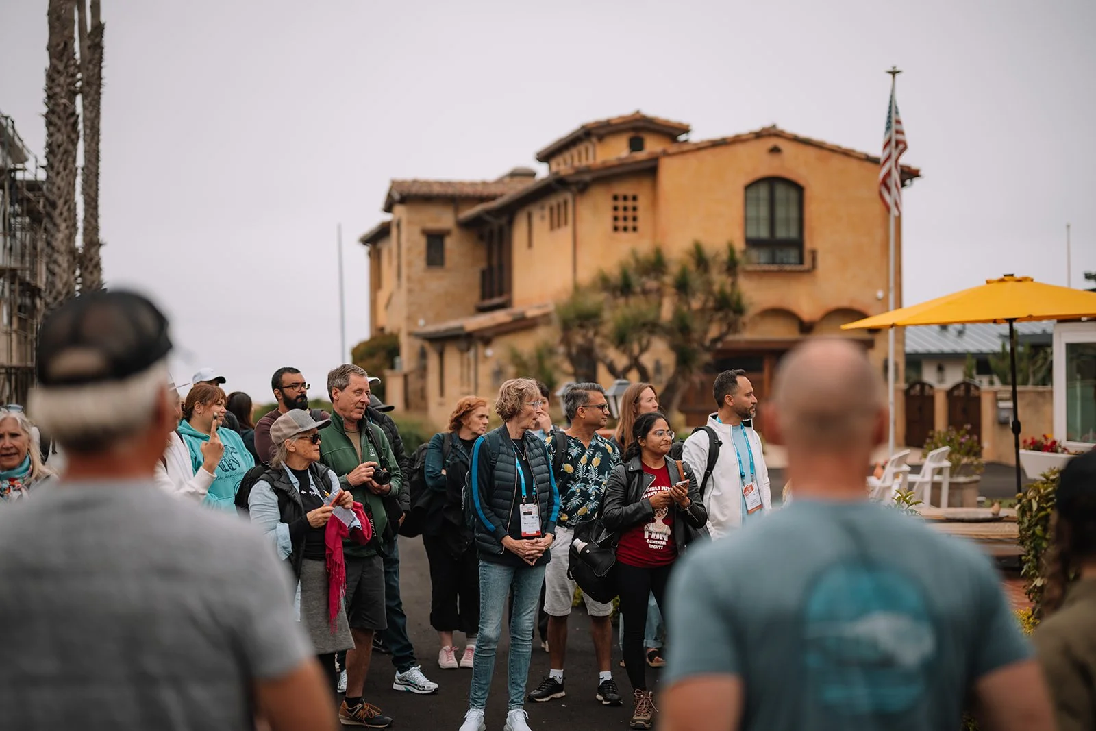 Tourists gathered outdoors in front of a yellow Spanish-style house with an American flag, overcast sky.