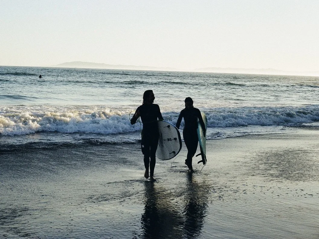 Two people walking out of the ocean holding surfboards on a sandy beach at sunset.