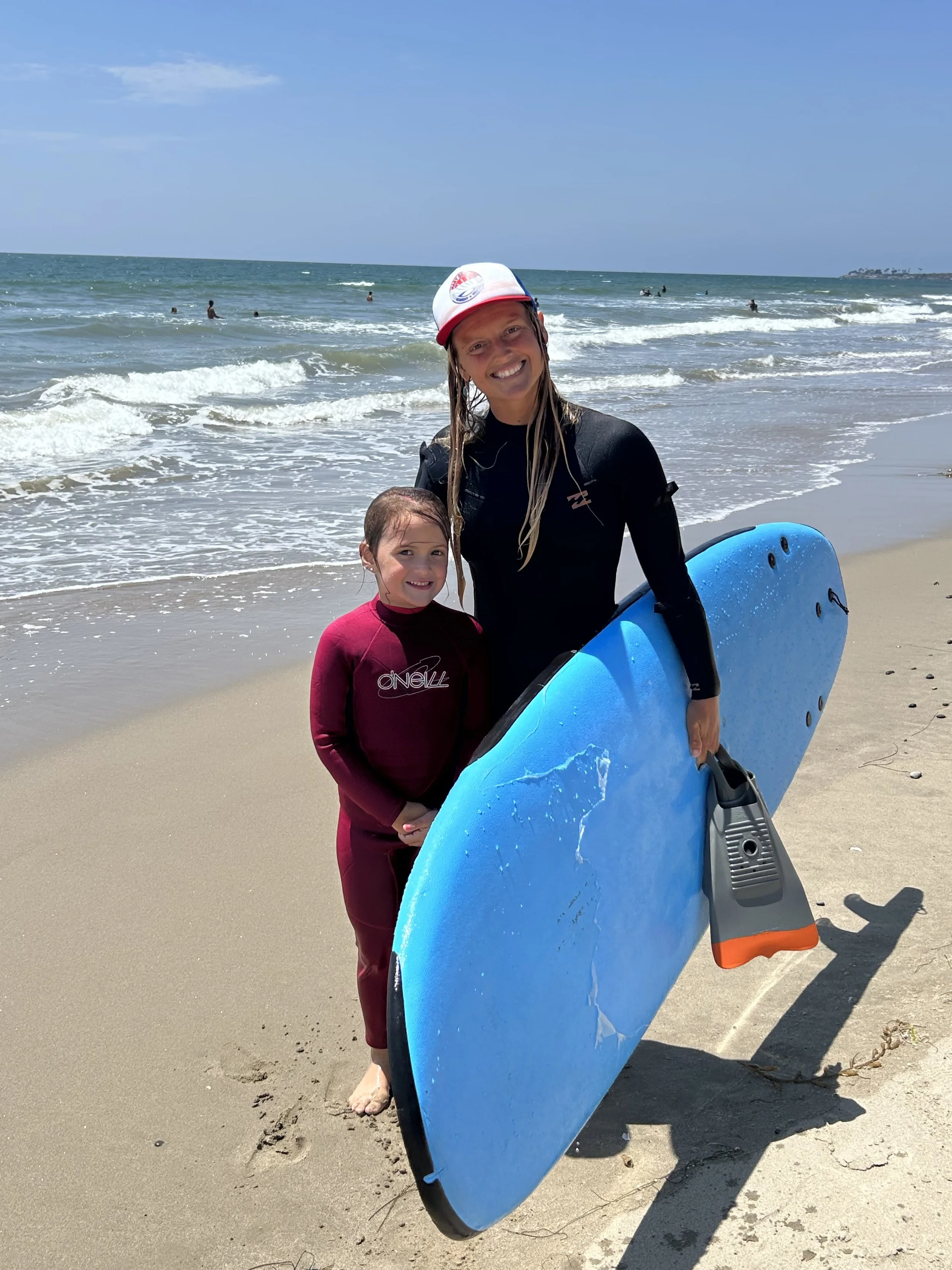A woman and a young girl standing on a beach, with the woman holding a blue surfboard, and both smiling, with the ocean and people swimming in the background.