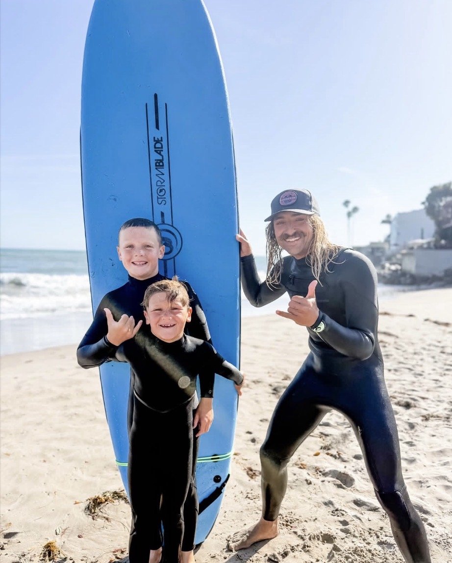 A man and two boys in wetsuits at the beach, with a surfboard, smiling and making shaka signs.