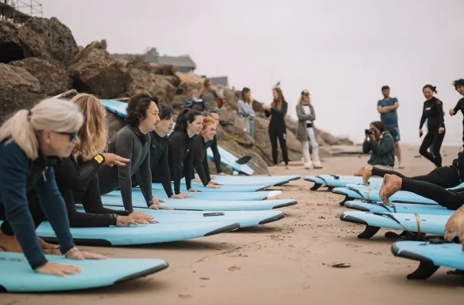 Group of people lying on surfboards on the beach, preparing for a surfing lesson, with others standing and watching nearby.