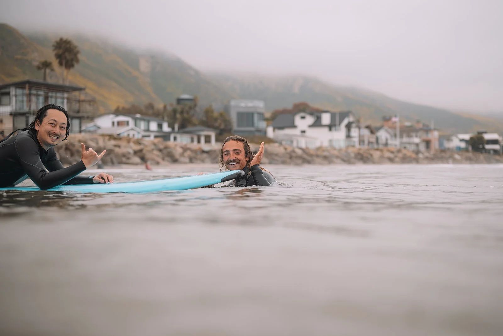 Two men in wetsuits smiling and making the shaka hand gesture while in the water near a surfboard, with a coastal town and mountains in the background on a foggy day.