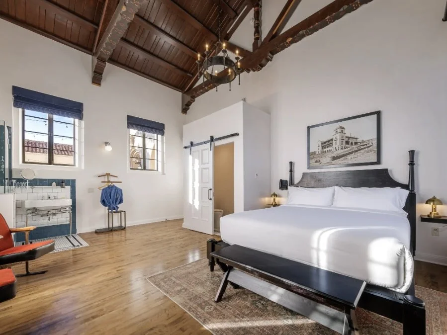 Interior of a spacious bedroom with a high, wooden ceiling, a large bed with white linens, artwork above the bed, modern bedside lamps, windows with blinds, a wooden floor, a vintage chandelier, and a sitting area with a red chair.