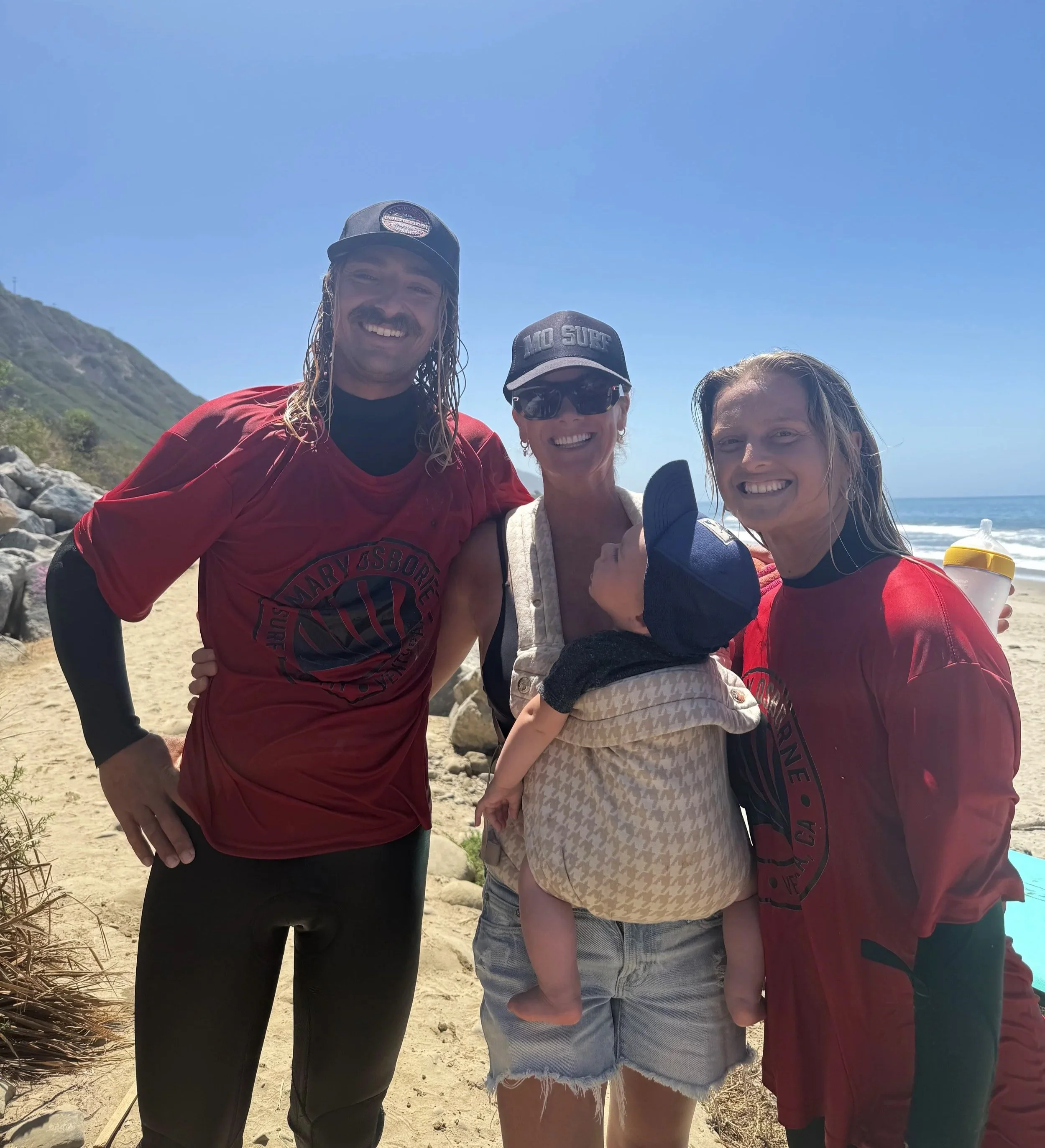 Three smiling people standing on a beach with ocean waves and sandy shore behind them. The first person on the left is a man with long, wet hair and a mustache, wearing a red shirt with black sleeves. The middle person is a woman with short hair, wearing sunglasses, a cap that says 'MO SURF', and a light-colored top. The third person on the right is a woman with long, wet hair, holding a yellow drink cup, wearing a red shirt.