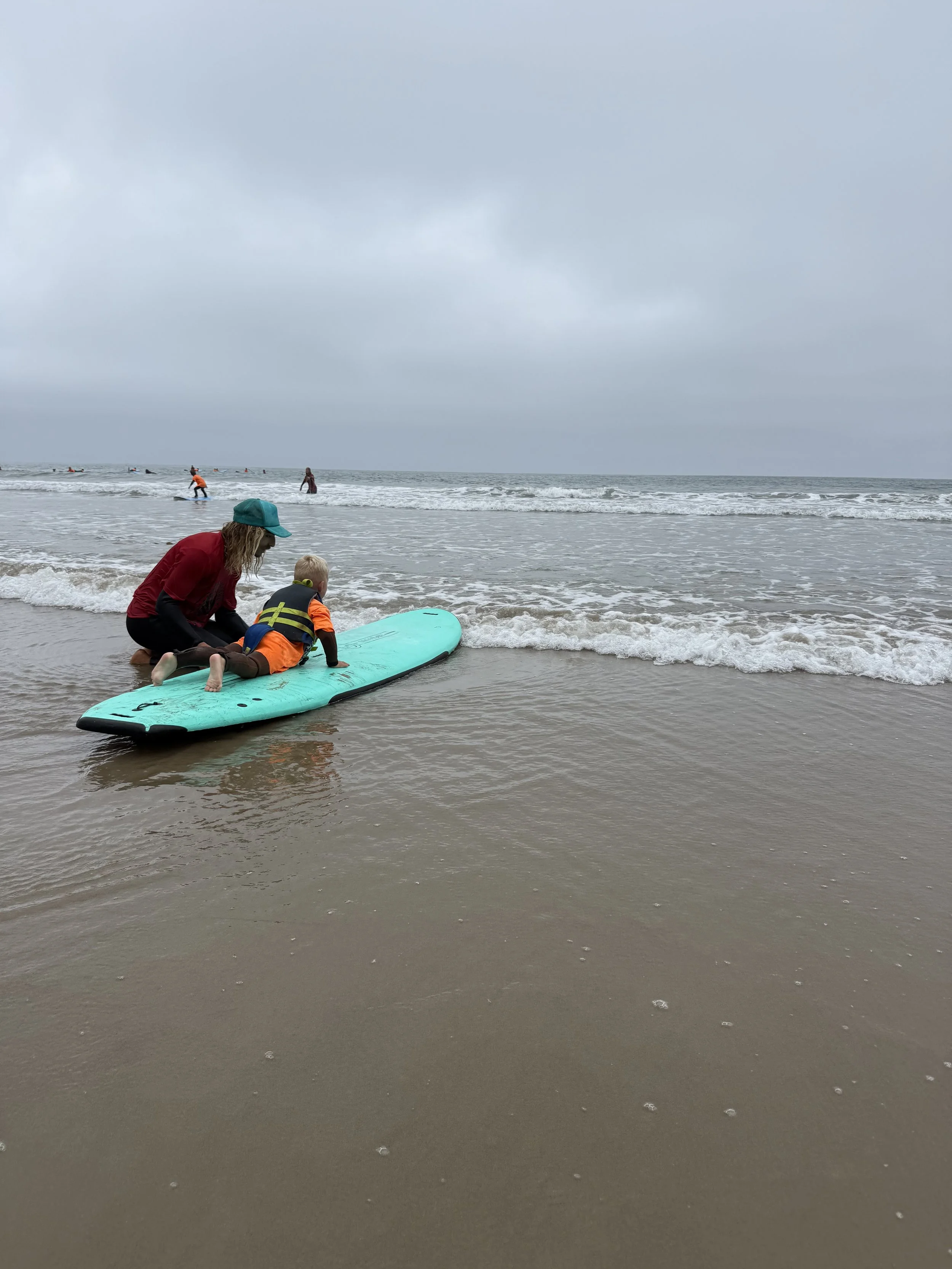 A woman and a young child in life jackets near the shoreline, with people surfing in the ocean under cloudy skies.