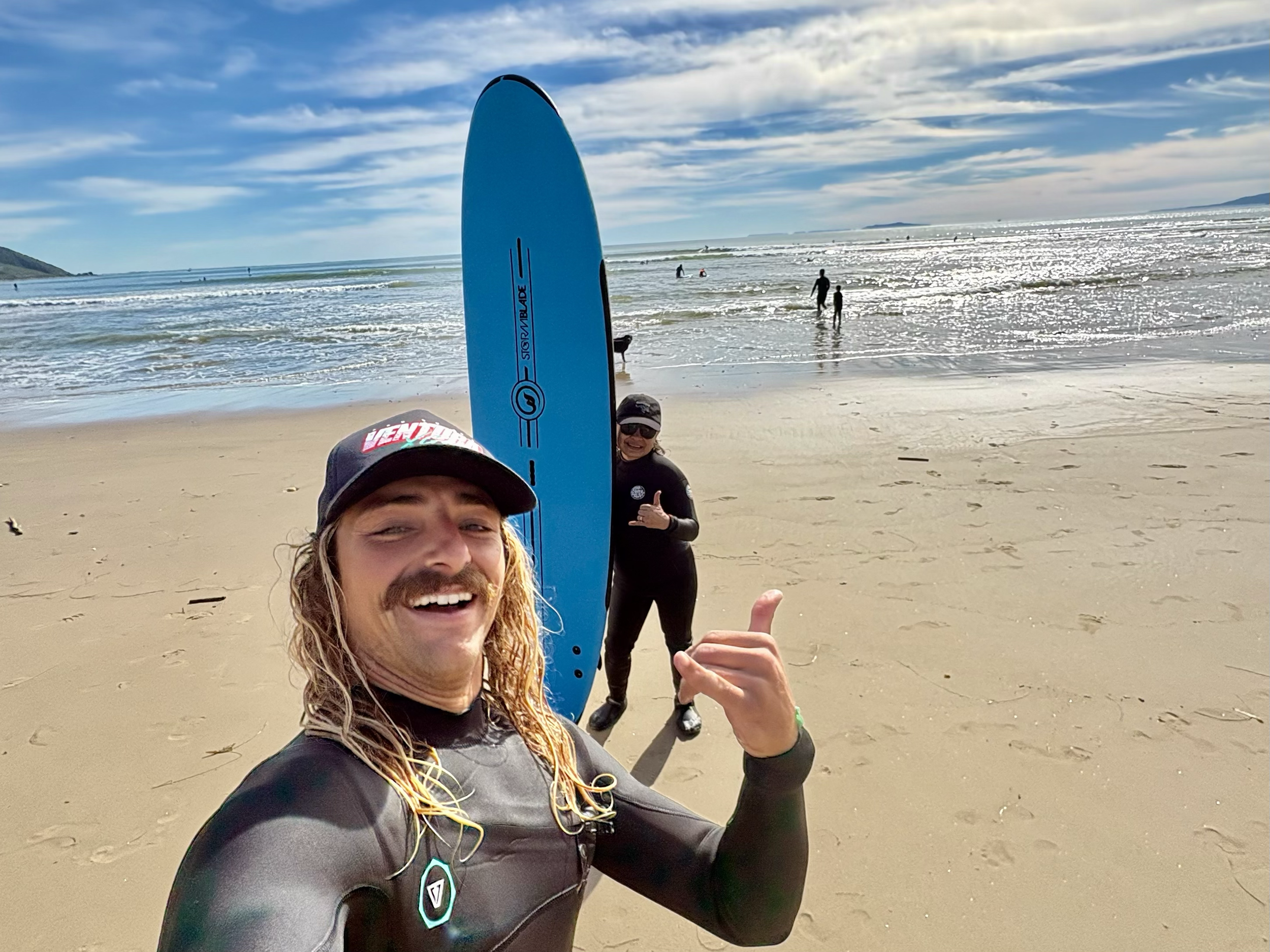 Group of people at the beach, some in wetsuits, with surfboards, on a sunny day with blue skies and ocean waves.