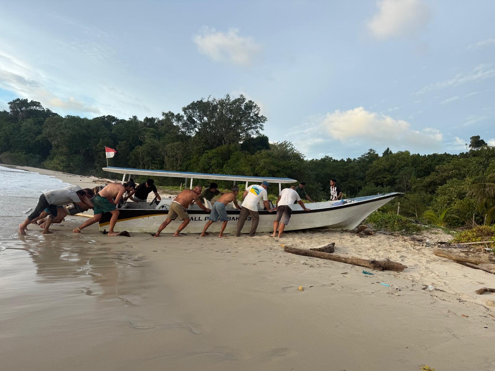 Ten men pushing a boat into the water on a sandy beach near a forested area with a cloudy sky overhead.