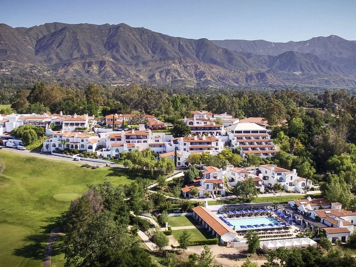 Aerial view of a resort with white buildings, a large swimming pool, and green landscaping, set against a backdrop of mountain range.