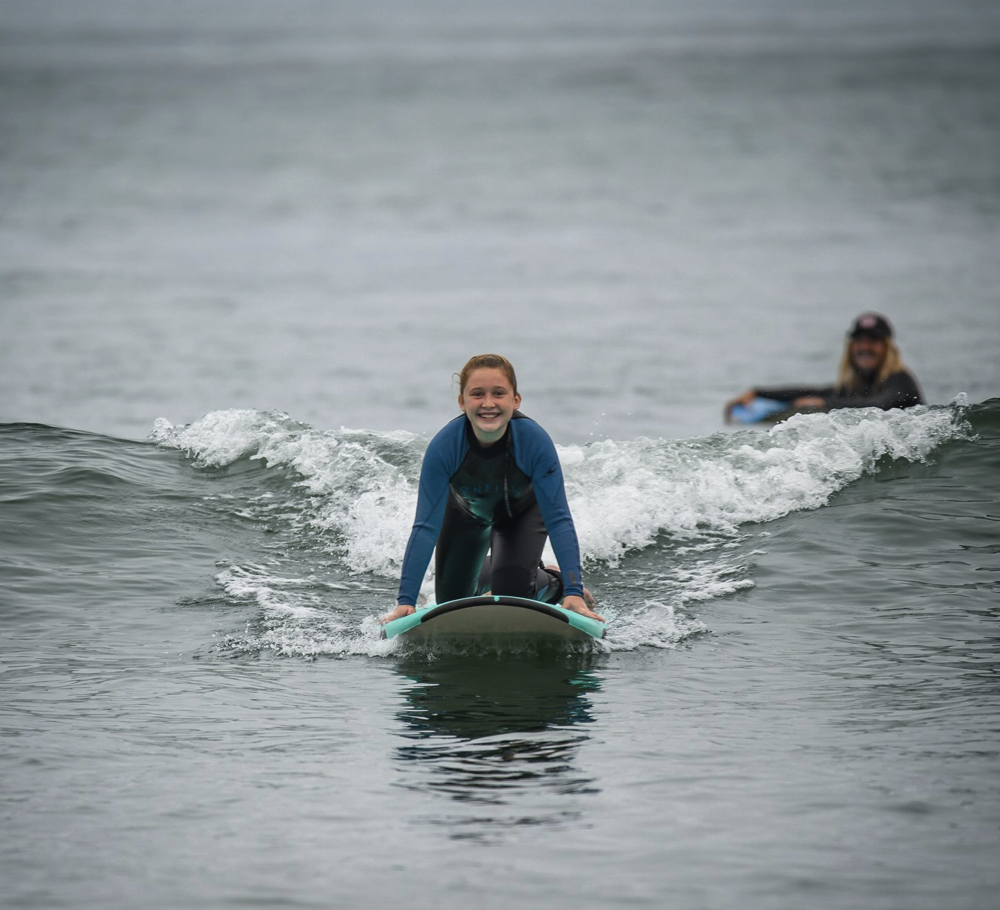 A young girl surfing on a wave with a smiling face, wearing a blue and black wetsuit, while a woman in the background watches.