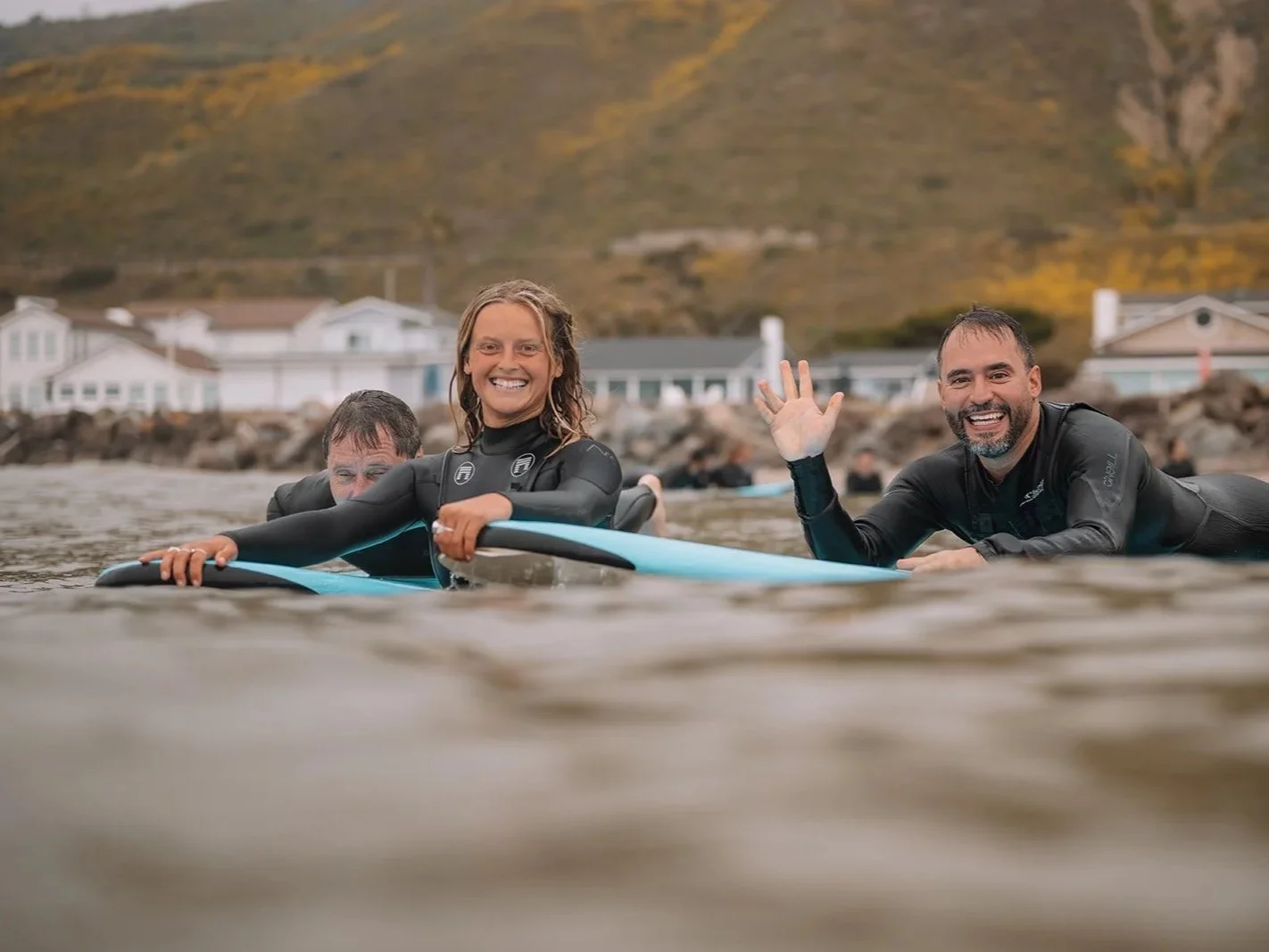 Three people in wetsuits on surfboards in the water, smiling and waving, with a background of houses and a hillside.