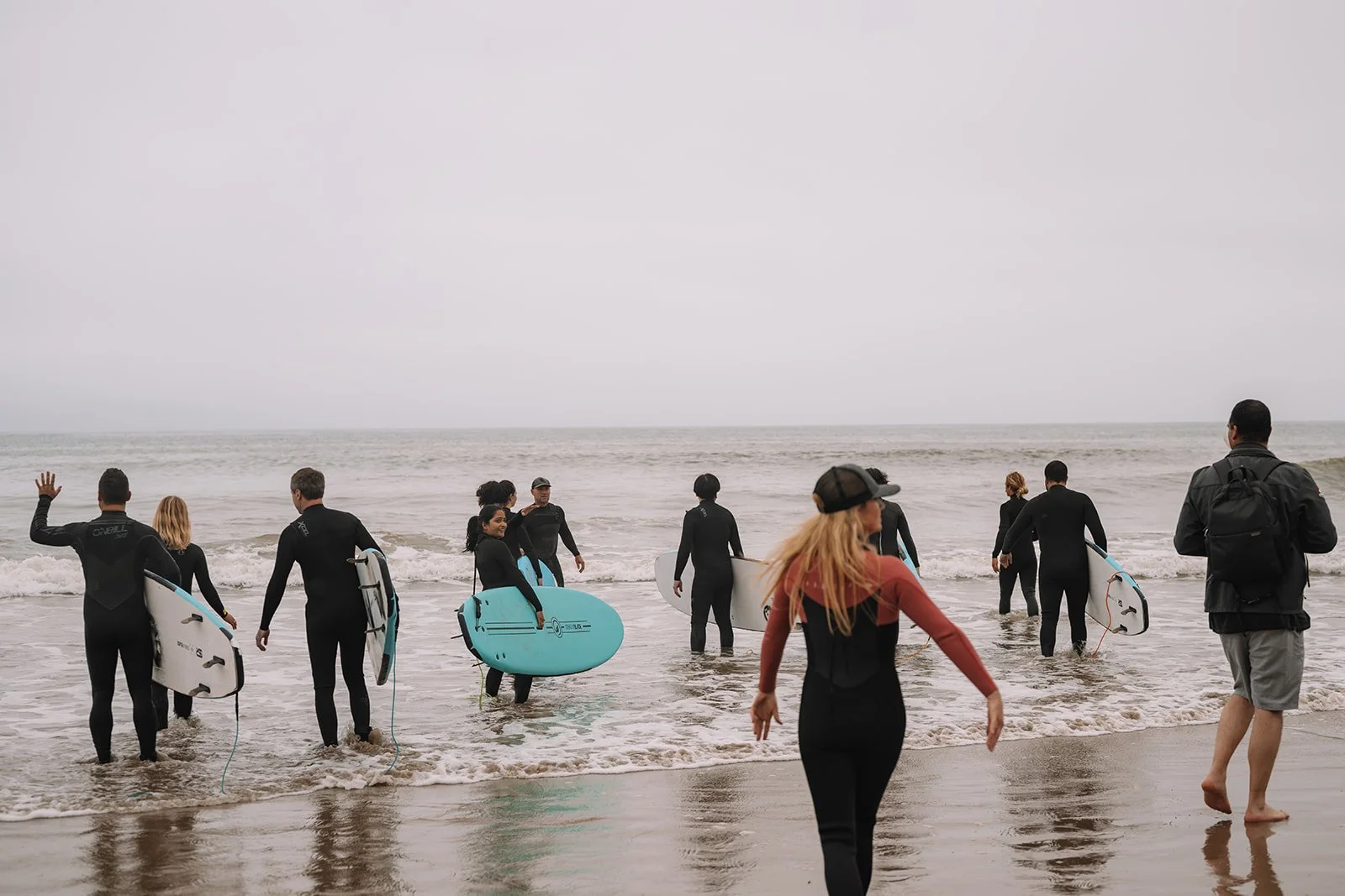 People at the beach with surfboards, some entering the water and some walking on the sand, under an overcast sky.