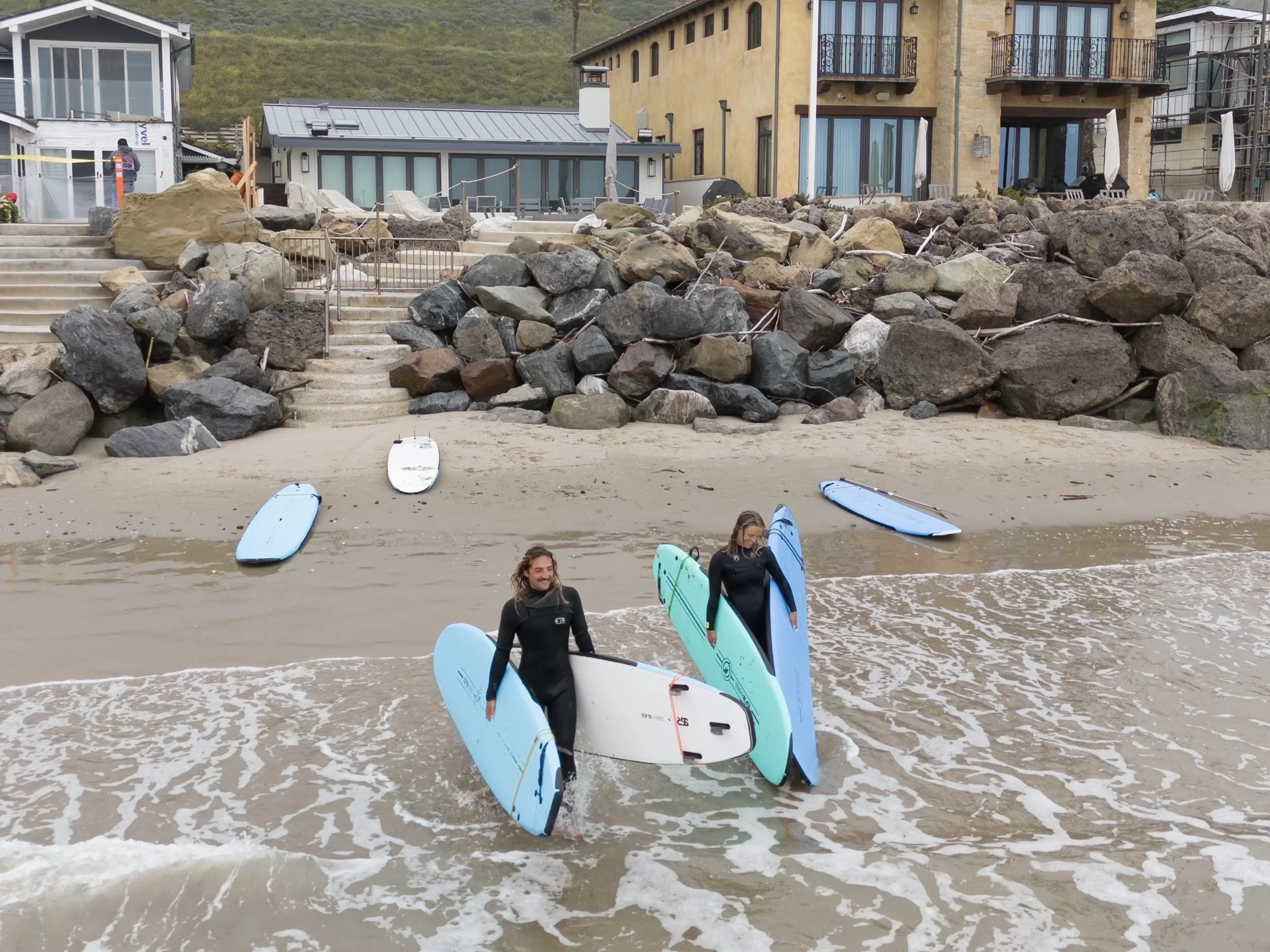 Two people in wetsuits holding surfboards standing in shallow ocean water with rocks and stairs leading up to a large building in the background.