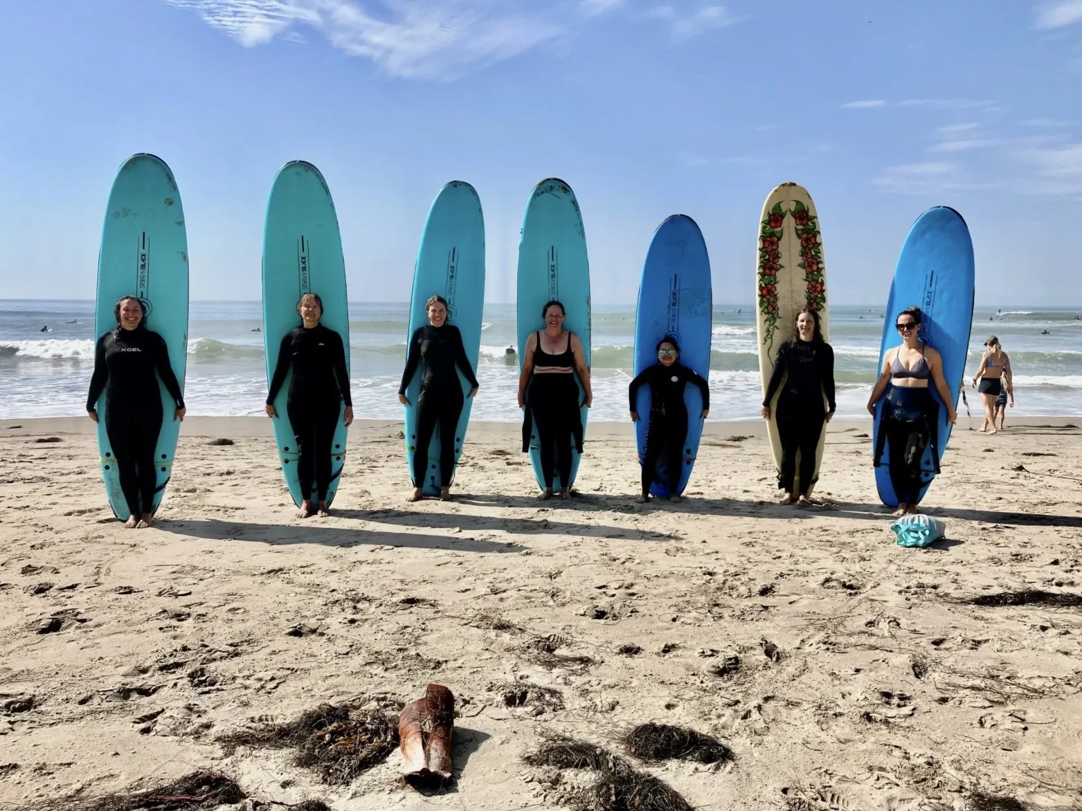 Group of seven women standing on a beach holding surfboards, with the ocean and people surfing in the background on a sunny day.