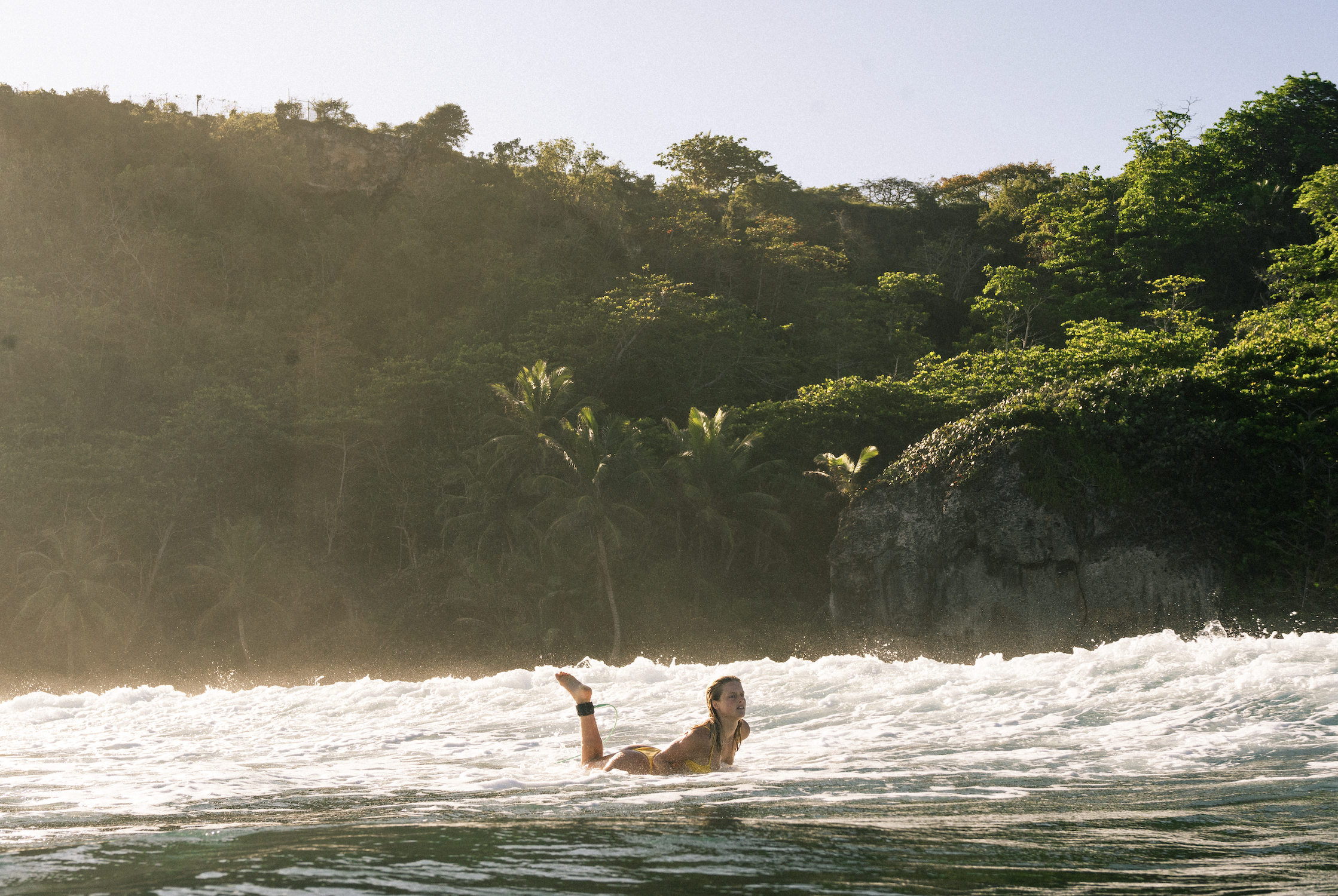 A girl in a yellow swimsuit is lying in the ocean surf near the shore with her hair wet, and a large lush green hillside with trees is visible in the background.