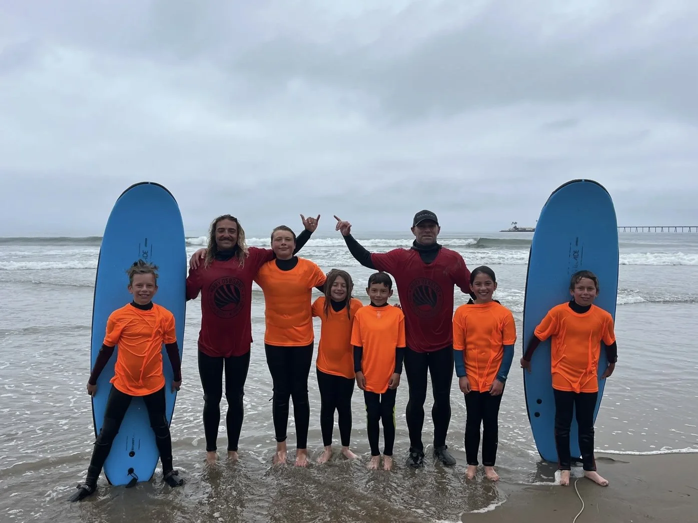 Group of children and two adults standing on a beach, holding surfboards. The children are wearing orange shirts, and the adults are in maroon shirts. They are smiling, with a cloudy sky and ocean waves in the background.