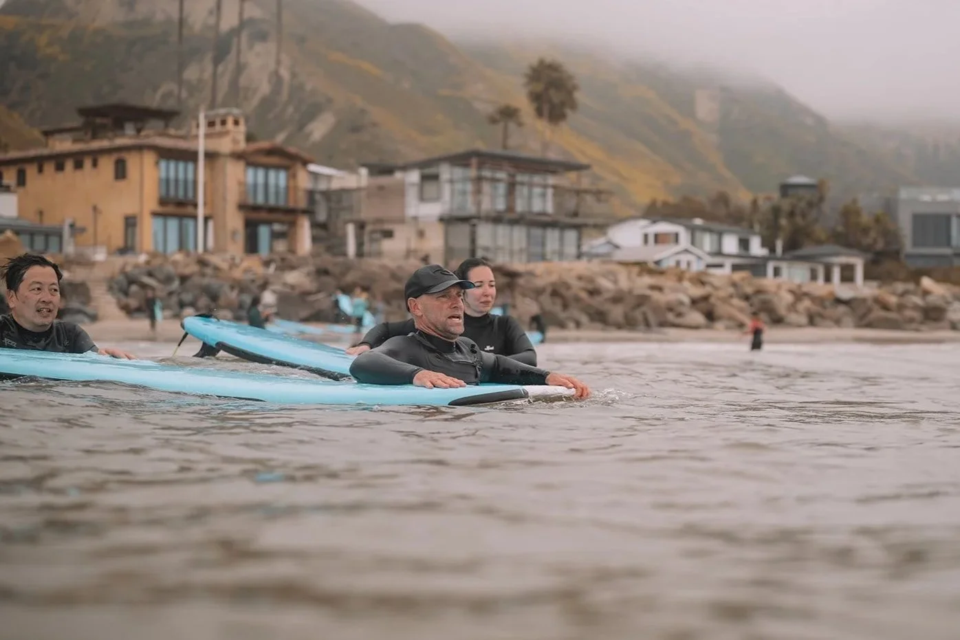 People in wetsuits practicing surfboarding in the ocean with a rocky shoreline and houses on hills in the background.