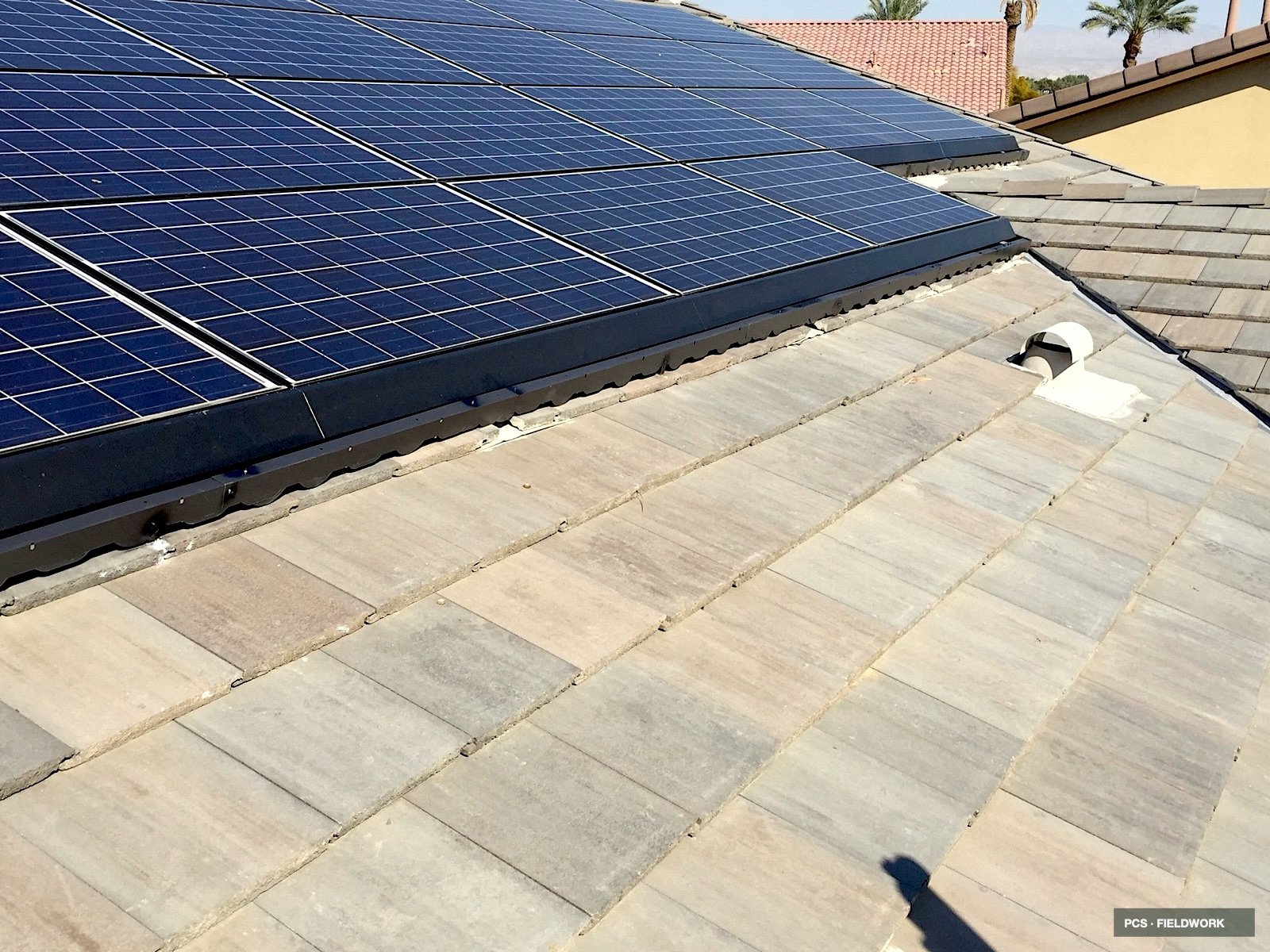 Solar panels installed on a rooftop with beige tiles, with other roofs and palm trees visible in the background.