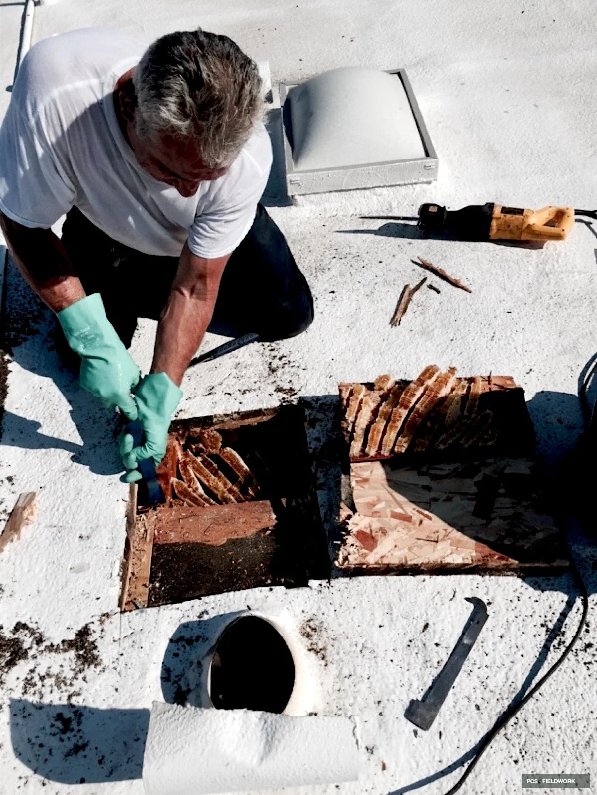 A person wearing a white shirt and green gloves working on a construction site, cutting brick with a reciprocating saw, with a hole in the white roofing material in the foreground.