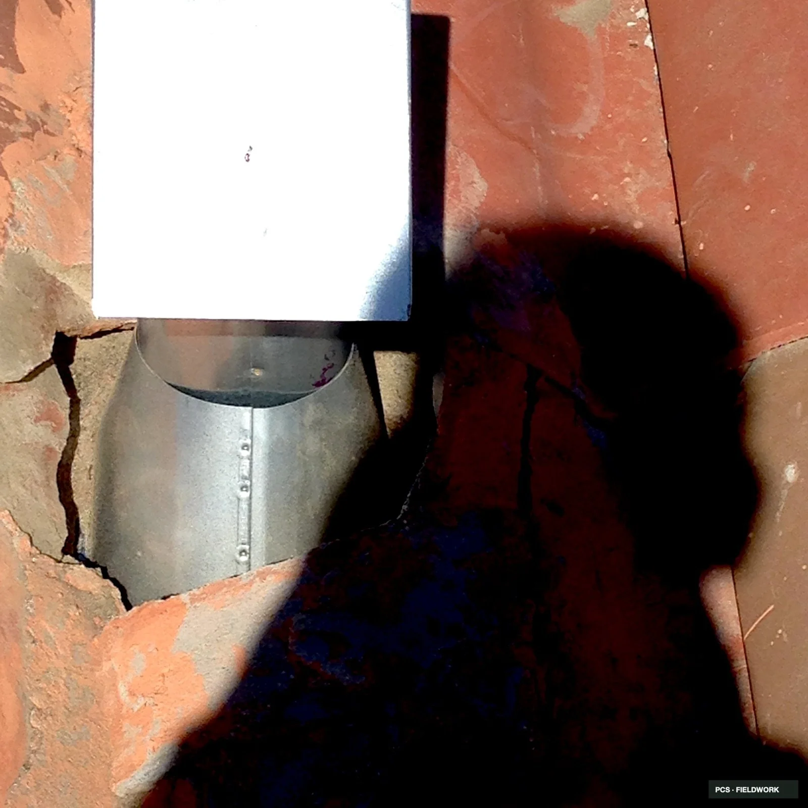 A bathroom with a toilet and metal tank on a brick wall, with part of a person standing nearby wearing a dark shirt and checkered shorts.