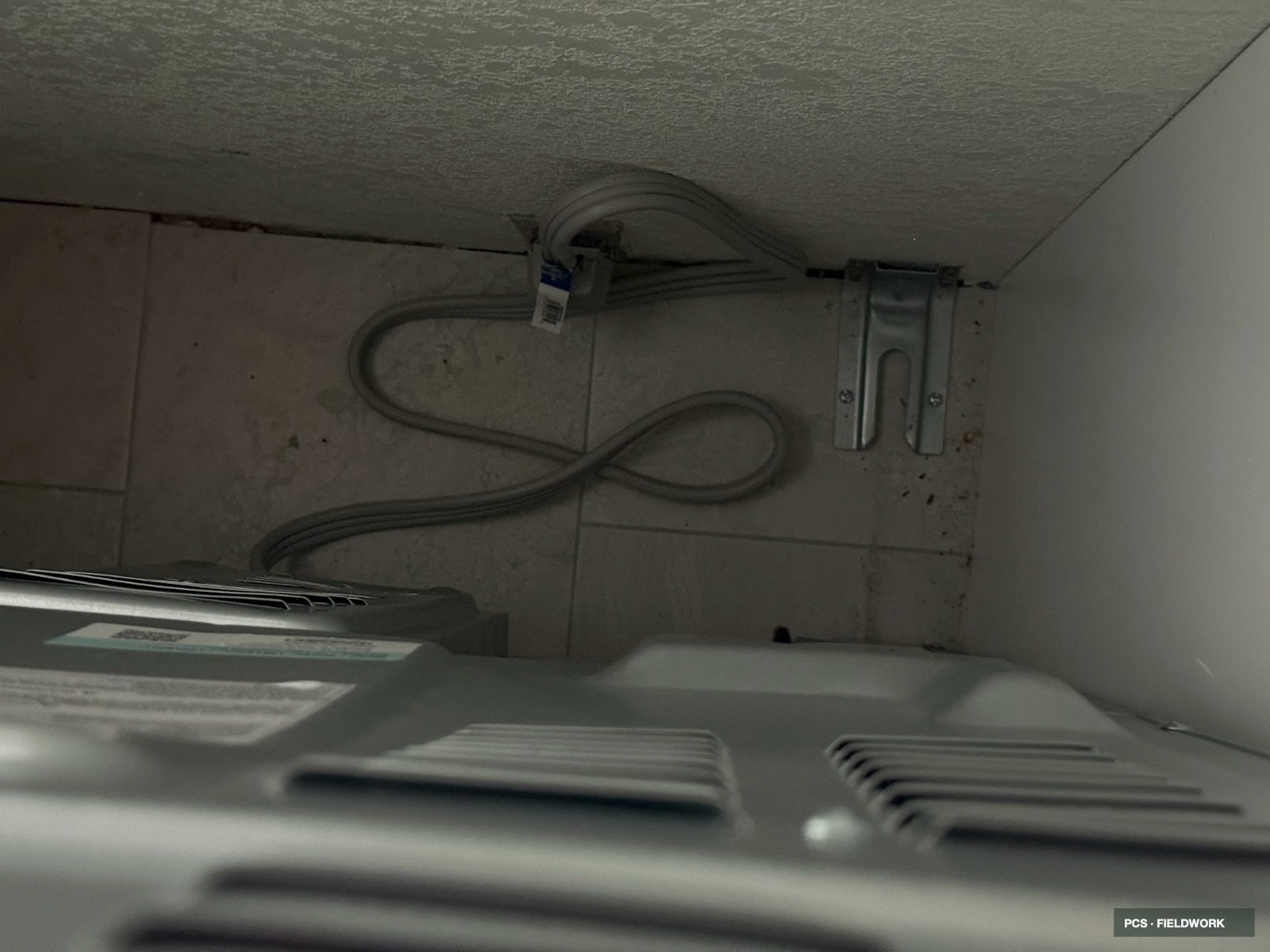 The image shows the floor of a laundry room with a drain pipe, a bend in the pipe, a metal bracket attached to the wall, and parts of a washing machine at the bottom of the photo.