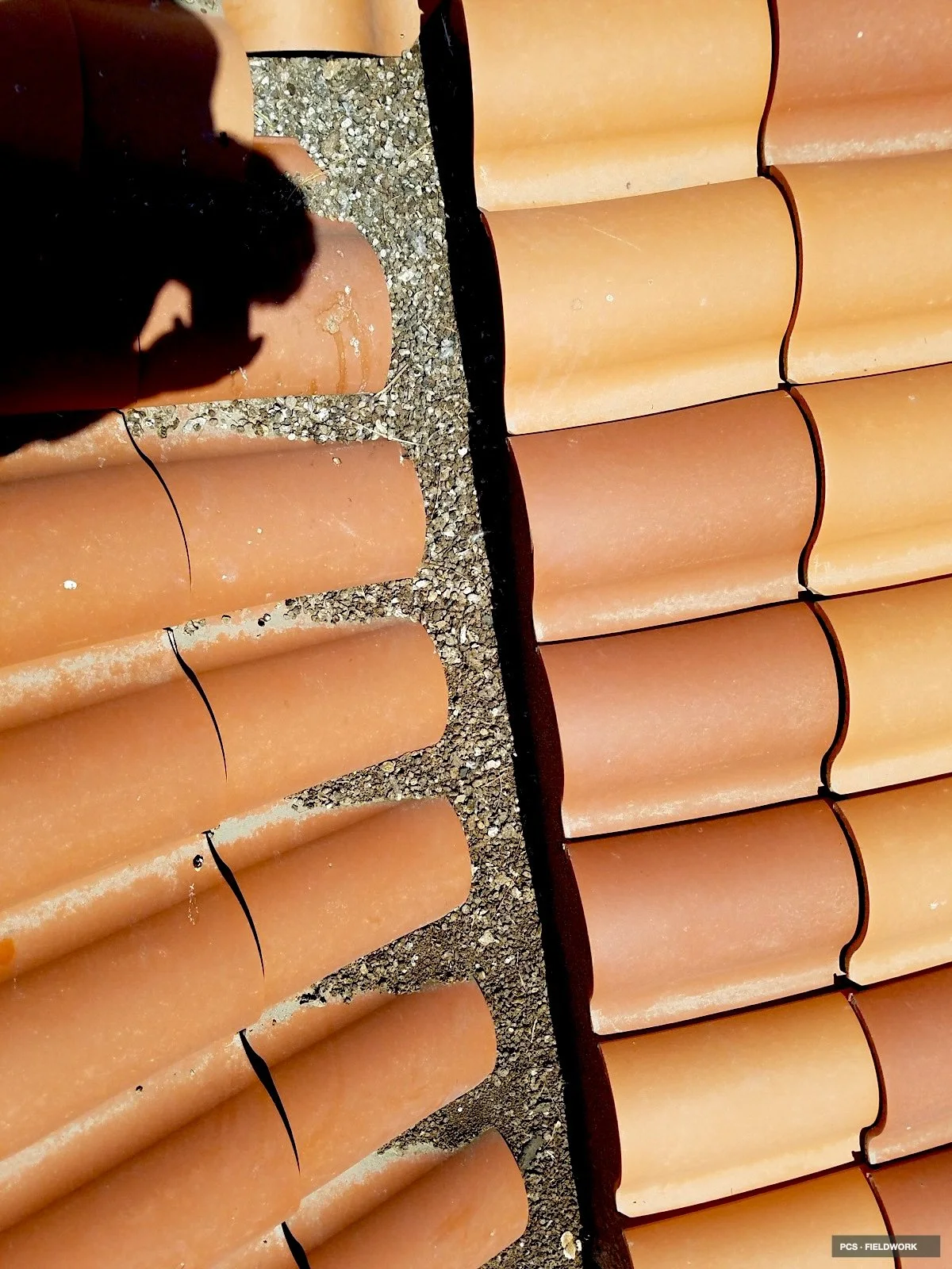 Close-up view of a terracotta-colored clay tile roof, showing individual curved tiles with black grouting in between, and some shadow and gravel visible underneath.