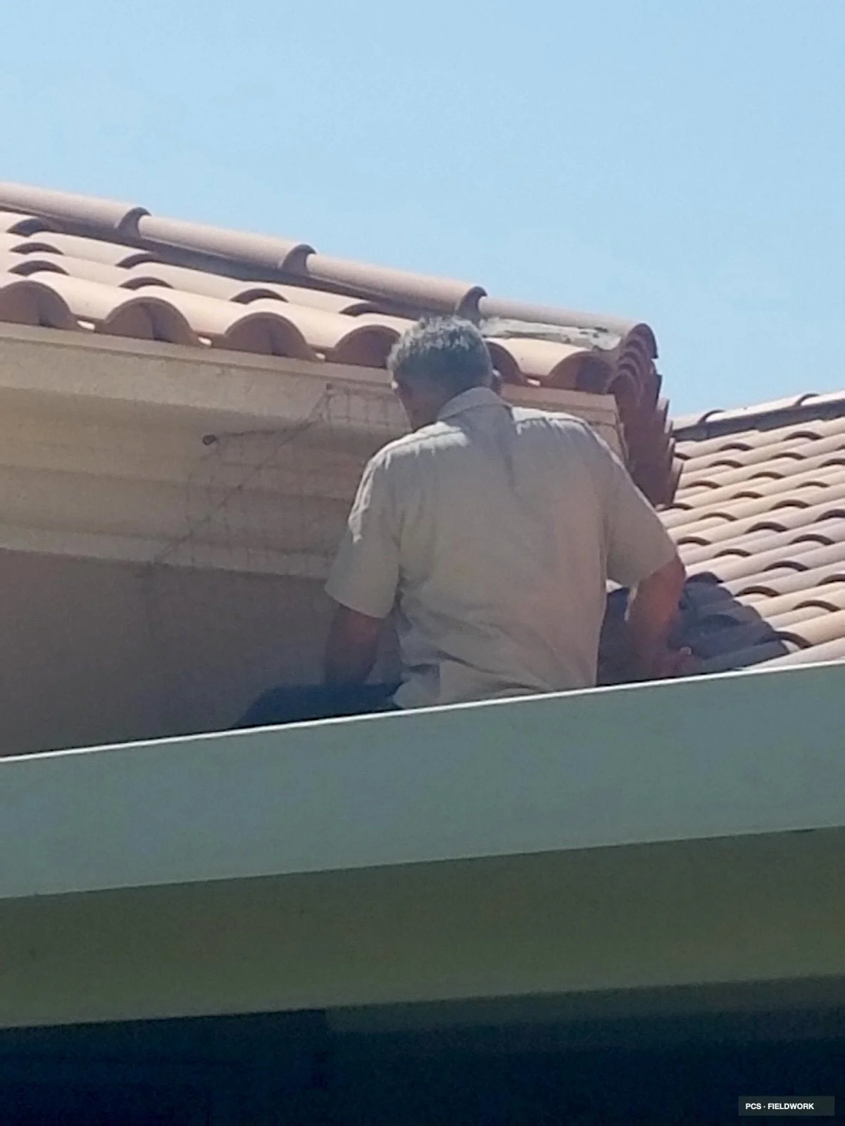 A man with gray hair, wearing a light-colored shirt, sitting on a roof with beige tiled roofing tiles, under a clear blue sky.