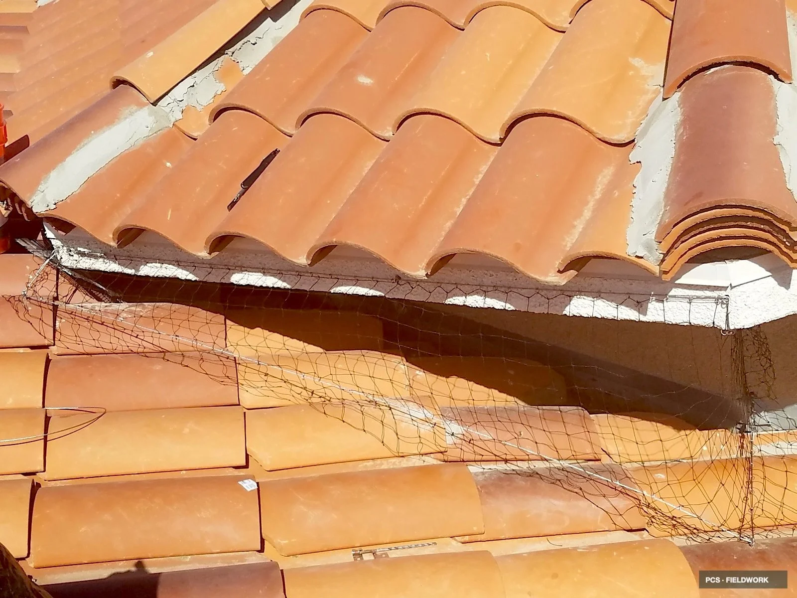 Stacked terracotta roof tiles stored under a net on the edge of a white wall, with some tiles damaged or broken.