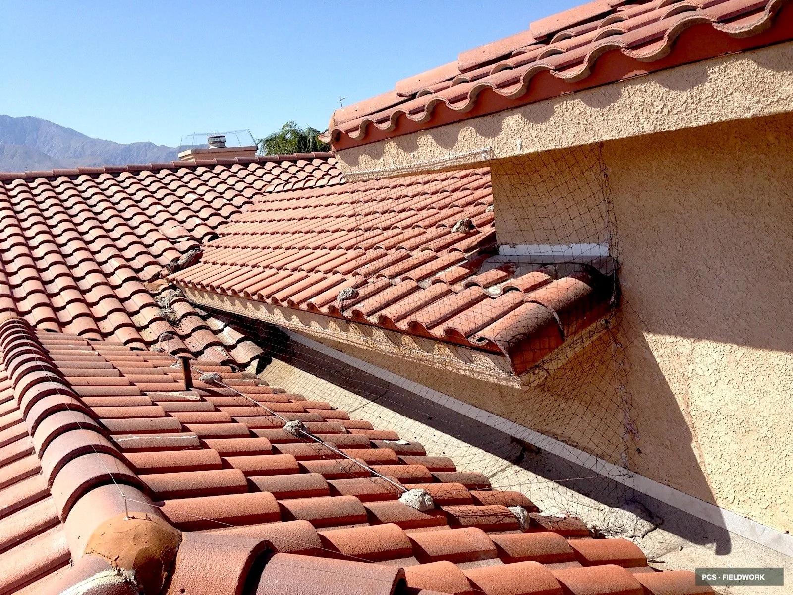 Roof with damaged and missing red clay tiles, some covered with a repair net, under a clear blue sky with mountains in the background.