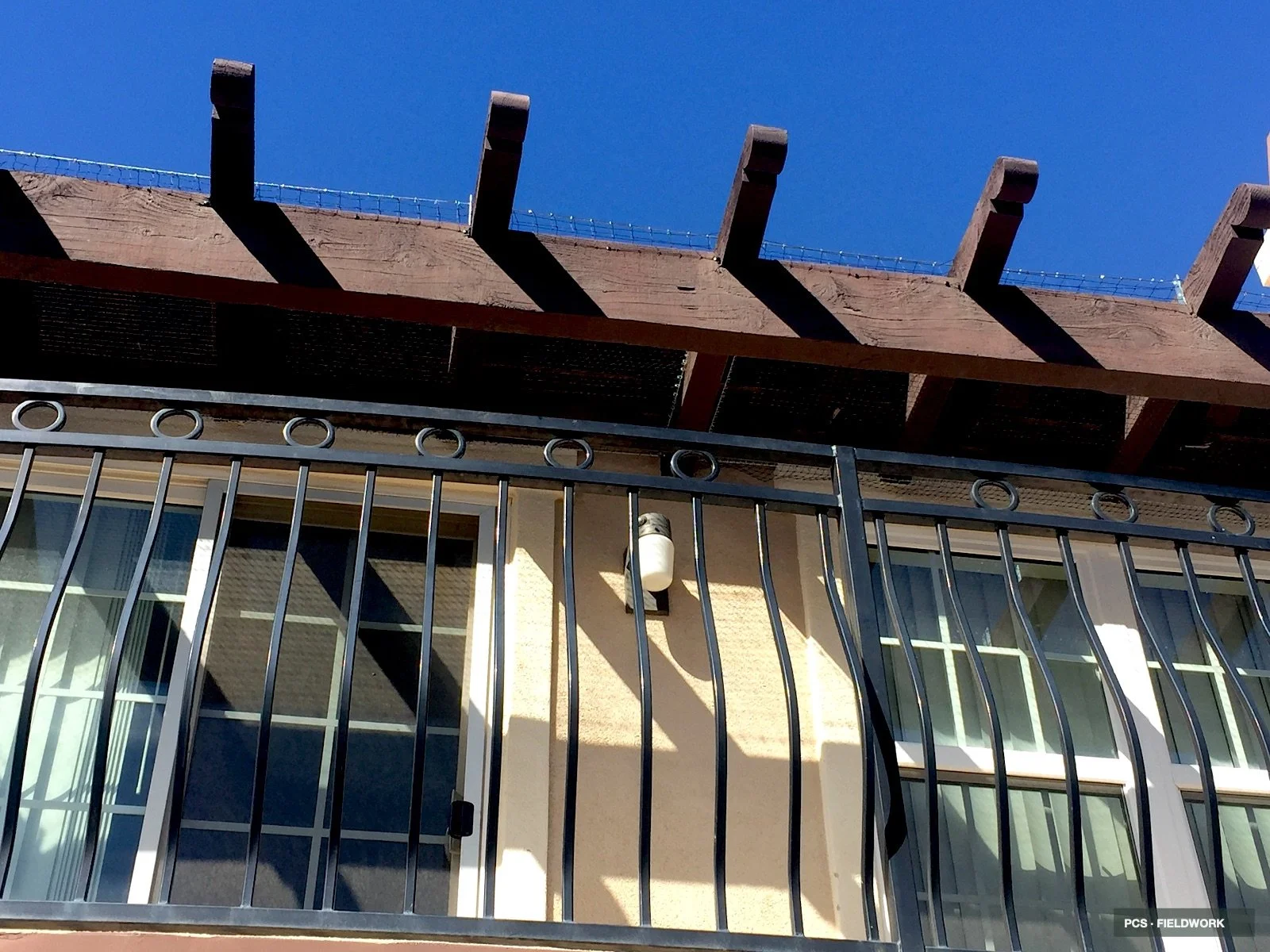 View of a residential balcony with a black metal railing in the foreground, a sliding glass door, exterior wall light fixture, and an overhanging wooden porch roof with large wooden beams and barbed wire along the top edge, under a clear blue sky.