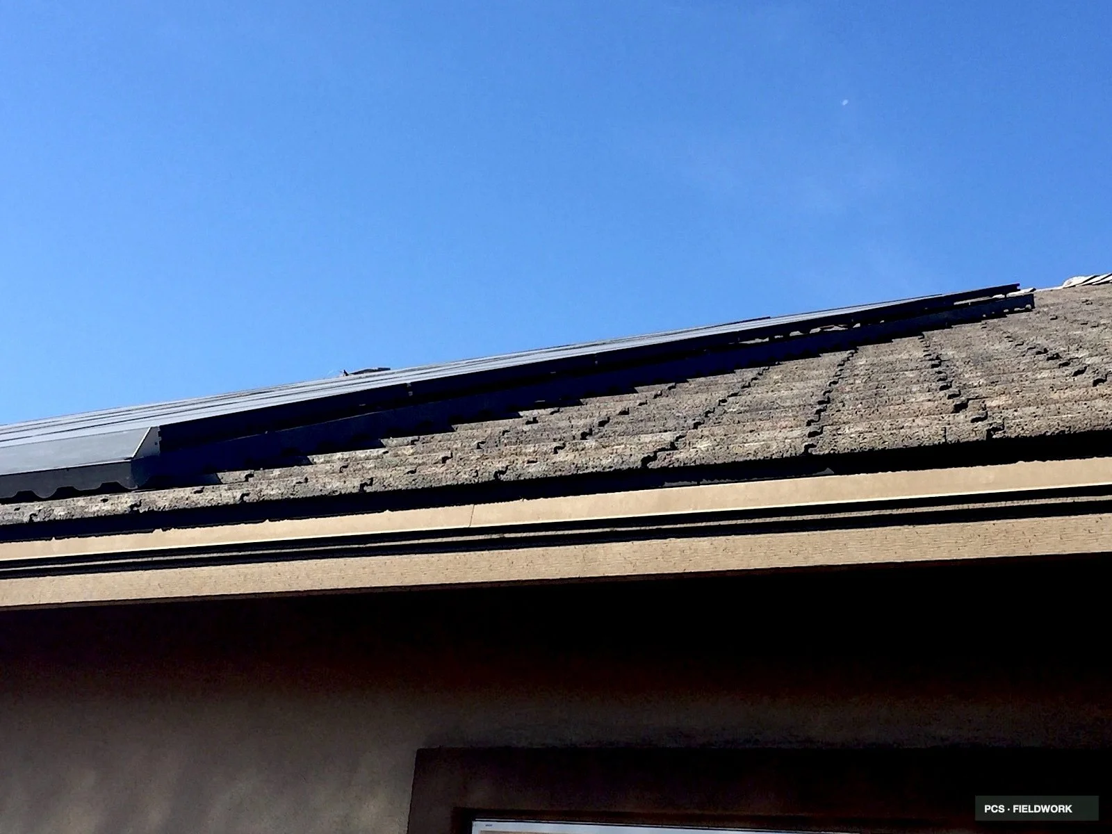 Close-up view of a house's roof with dark shingles and a metal gutter, against a blue sky.