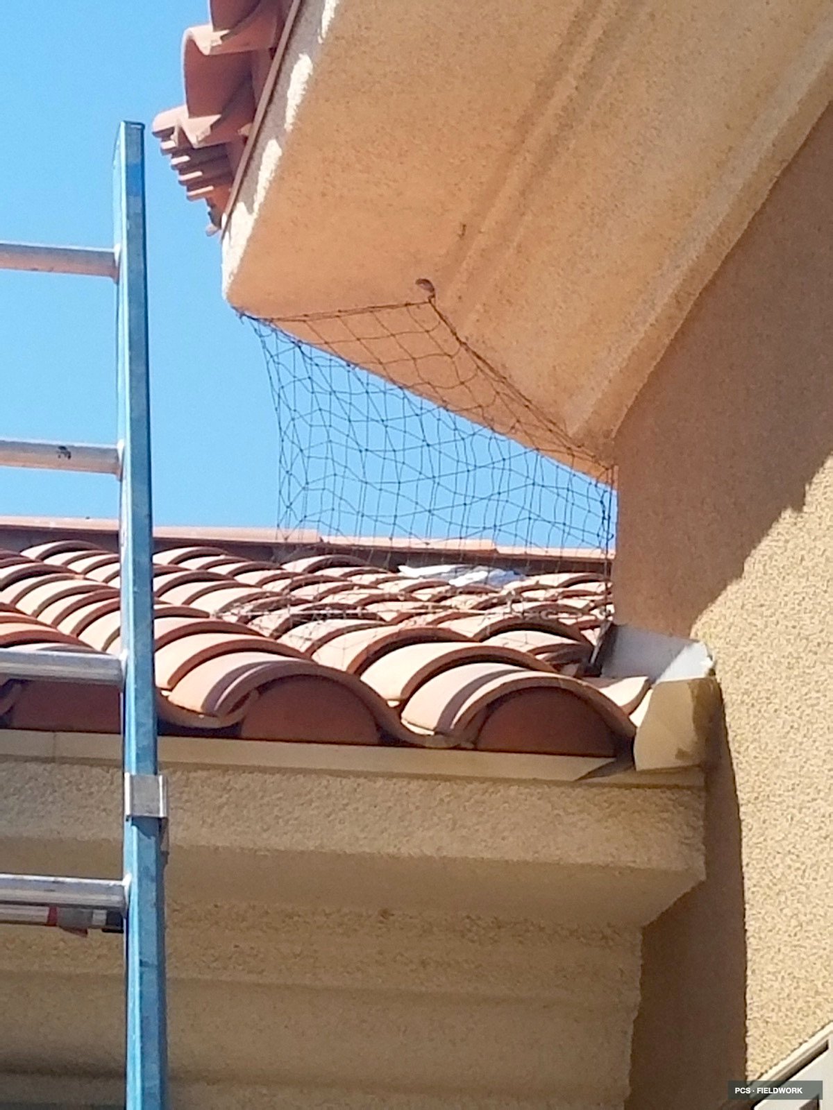 Outside view of a building with clay roof tiles, a balcony with solid beige walls, a metal ladder, and a wire safety net on the balcony's edge, under a clear blue sky.