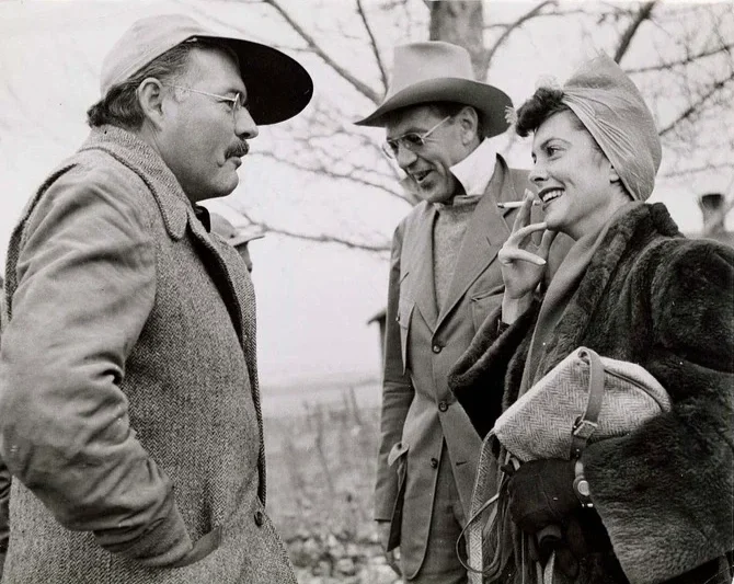 Three people standing outdoors and talking, two men and one woman, dressed warmly with hats, in a black-and-white photograph. Gary Cooper and Hemingway.