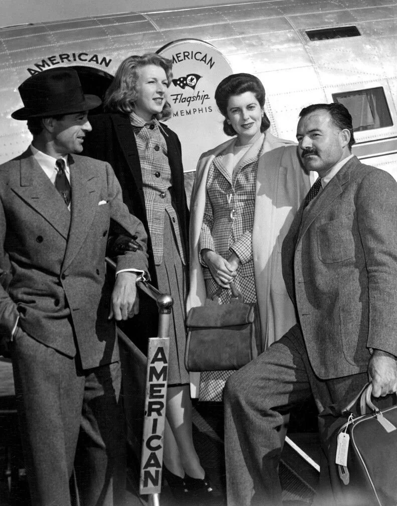 Four people standing in front of an airplane with the sign 'American' visible, at an airport or airfield. Gary Cooper. Ernest Hemingway.