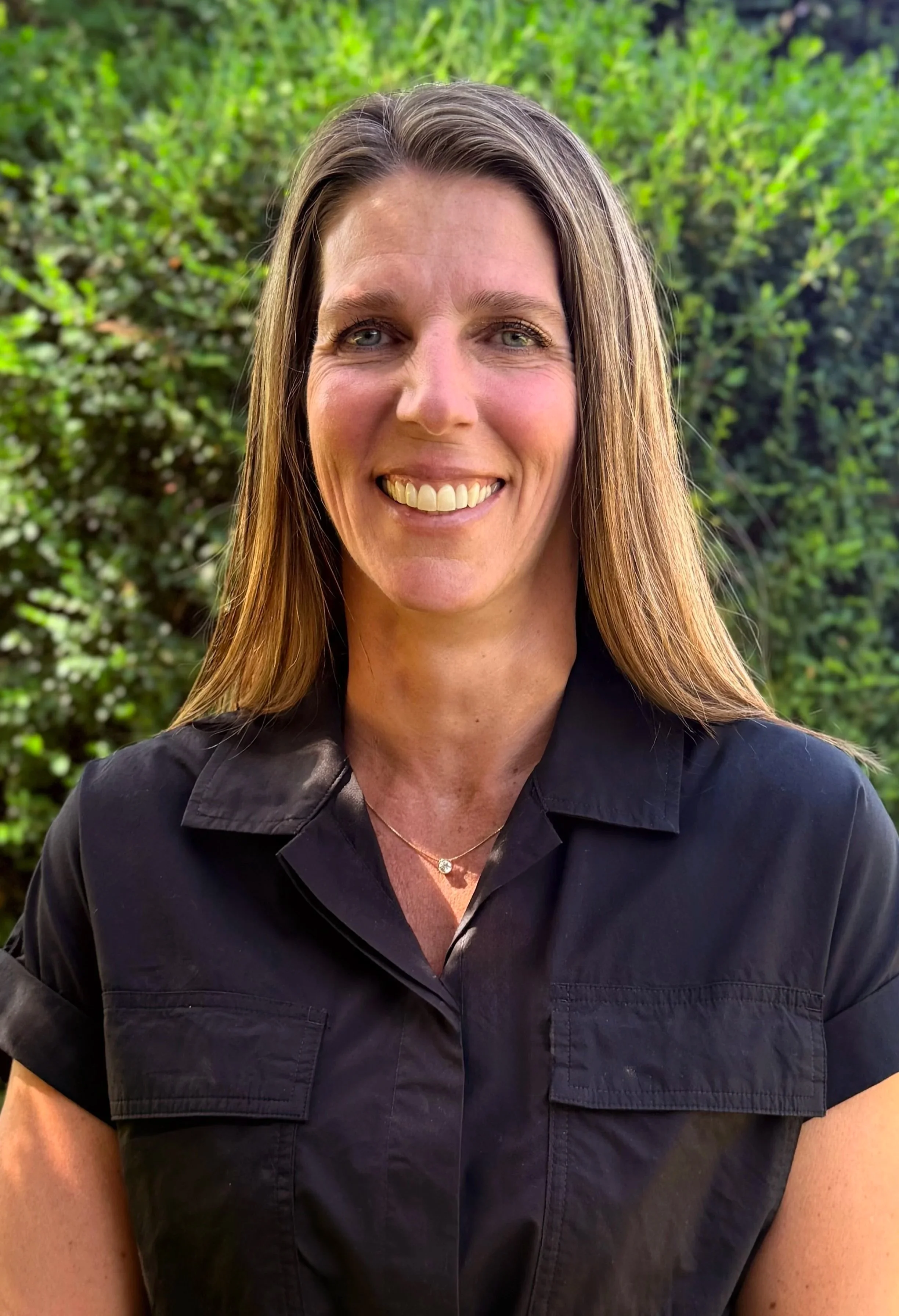 Close-up of a woman with long, brown hair and blue eyes, smiling, outdoors with green foliage in the background, wearing a black shirt and a delicate necklace.