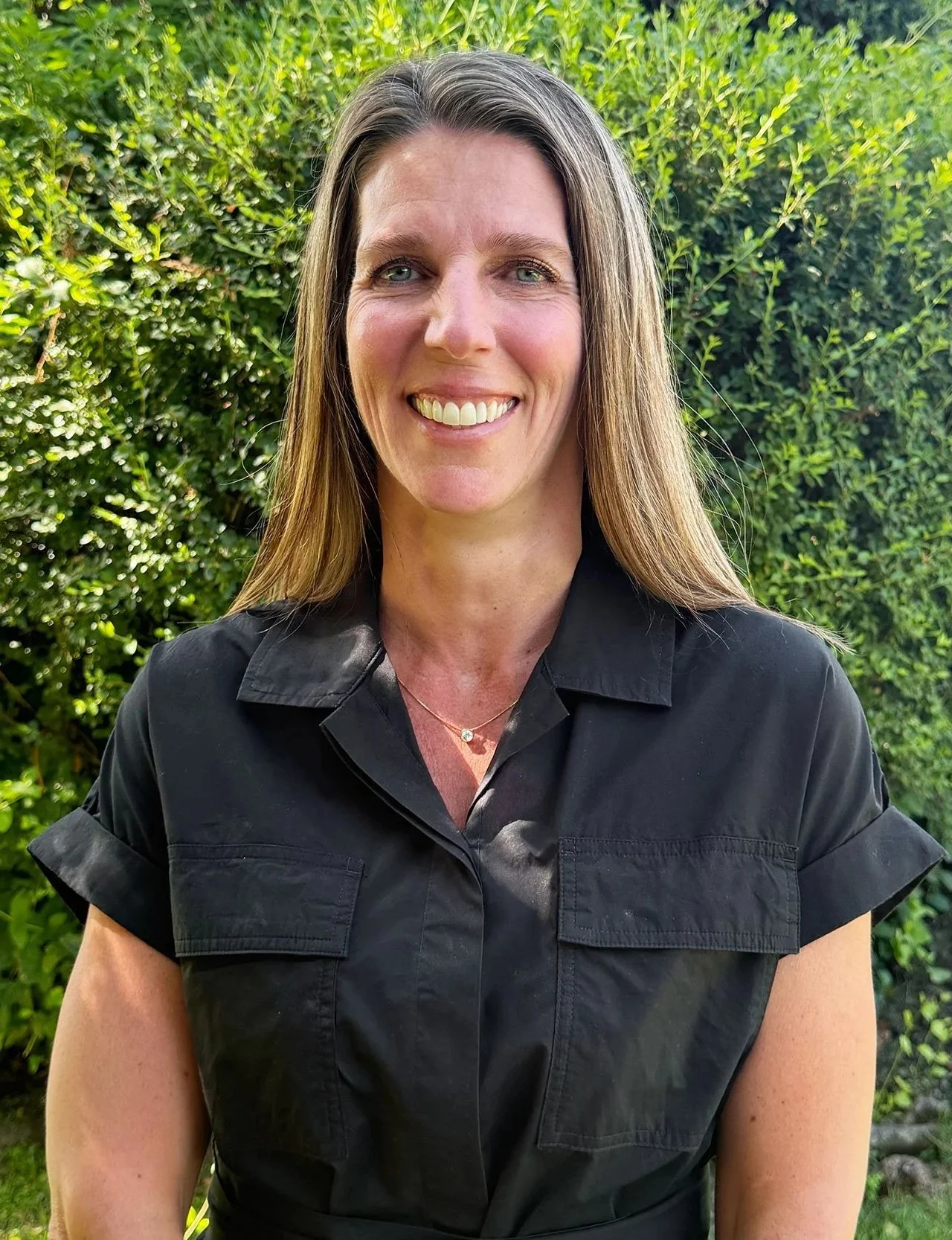 A woman with long brown hair, wearing a black short-sleeved shirt, smiling, standing outdoors in front of green bushes.